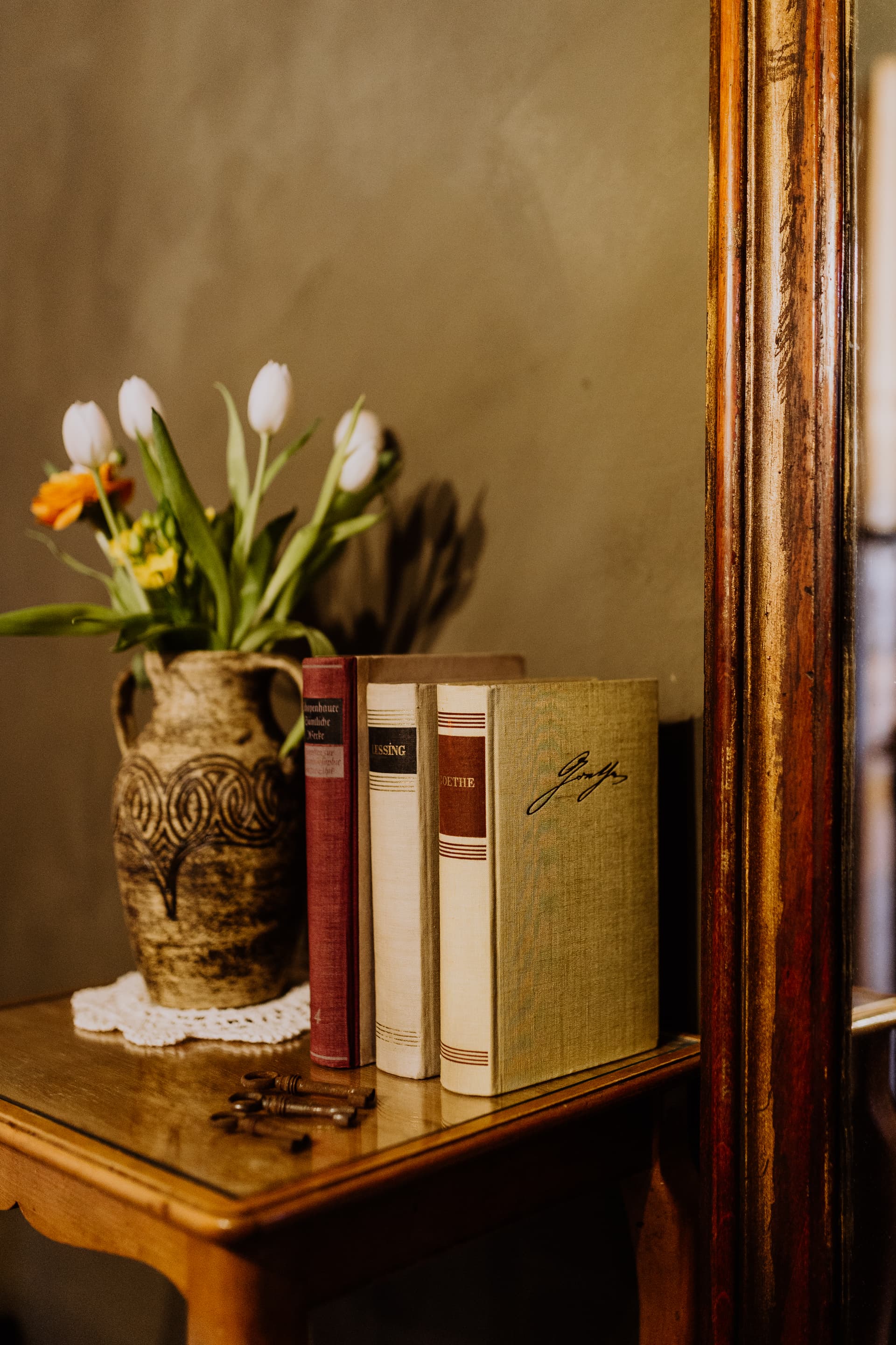 Frische Tulpen im Steingutkrug neben antiken Büchern und alten Schlüsseln vor dem Goldspiegel